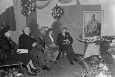 Aandachtig luistert de familie Regouin naar een spreker; naast hen een portret van oprichter M.B. Regouin (?). Cuijk, 1950