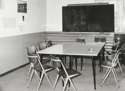 Krijtbord met omrekentabellen, een voetbalposter en asbakken op tafel in ruimte voor Spaanse arbeiders in Schijndel (foto: beeldcollectie Jansen de Wit, collectie BHIC nr. 1906-001535)